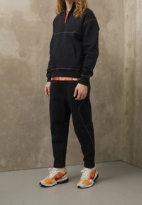 Sudadera negra con cremallera y costuras en contraste, con bolsillo canguro, combinada con pantalones jogger negros. Calzado con detalles en naranja y blanco.