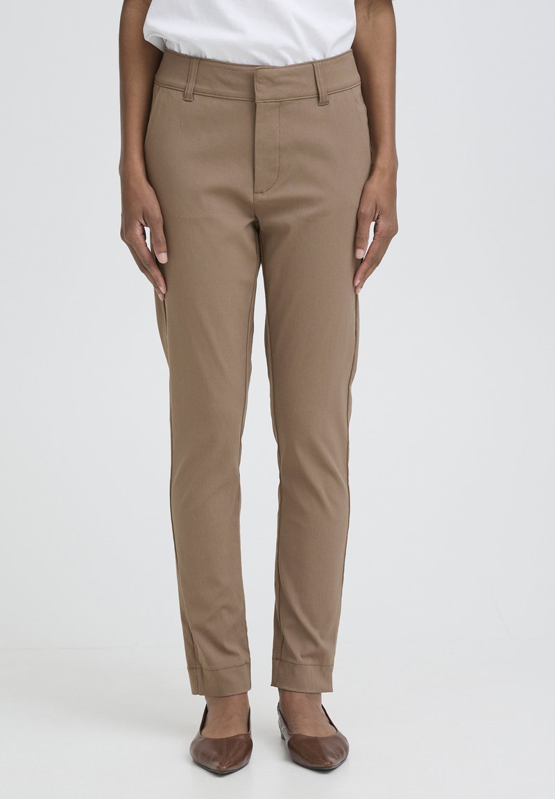 Personne debout, portant un pantalon droit beige, une chemise blanche rentrée et des ballerines marron devant un fond uni clair.