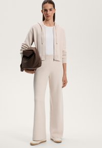 Felpa beige chiaro con cordini, top bianco, pantaloni svasati beige chiaro e sneaker beige. Tiene una borsa marrone scuro. Texture liscia.