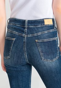 Vaqueros de mezclilla azul oscuro con un diseño ajustado, que cuentan con dos bolsillos traseros con costuras sutiles y una etiqueta marrón en la cinturilla.