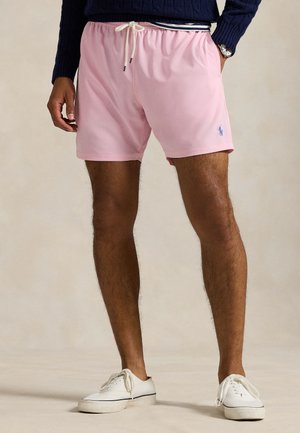 Mees, kes kannab roosat paelaga lühikesi pükse sinise tikandi ja logoga, tumelasinist kampsunit, valgeid tossusid ja randmekella neutraalse taustaga.