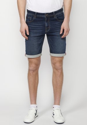 Shorts en denim bleu foncé avec des poignets roulés, présentant cinq poches et des coutures en contraste. Portés avec des baskets blanches et des chaussettes de cheville.