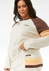 Sudadera de color beige claro con mangas raglán marrones, que presenta un bolsillo de rayas horizontales en colores pastel. Logo de "Rip Curl" en el pecho.