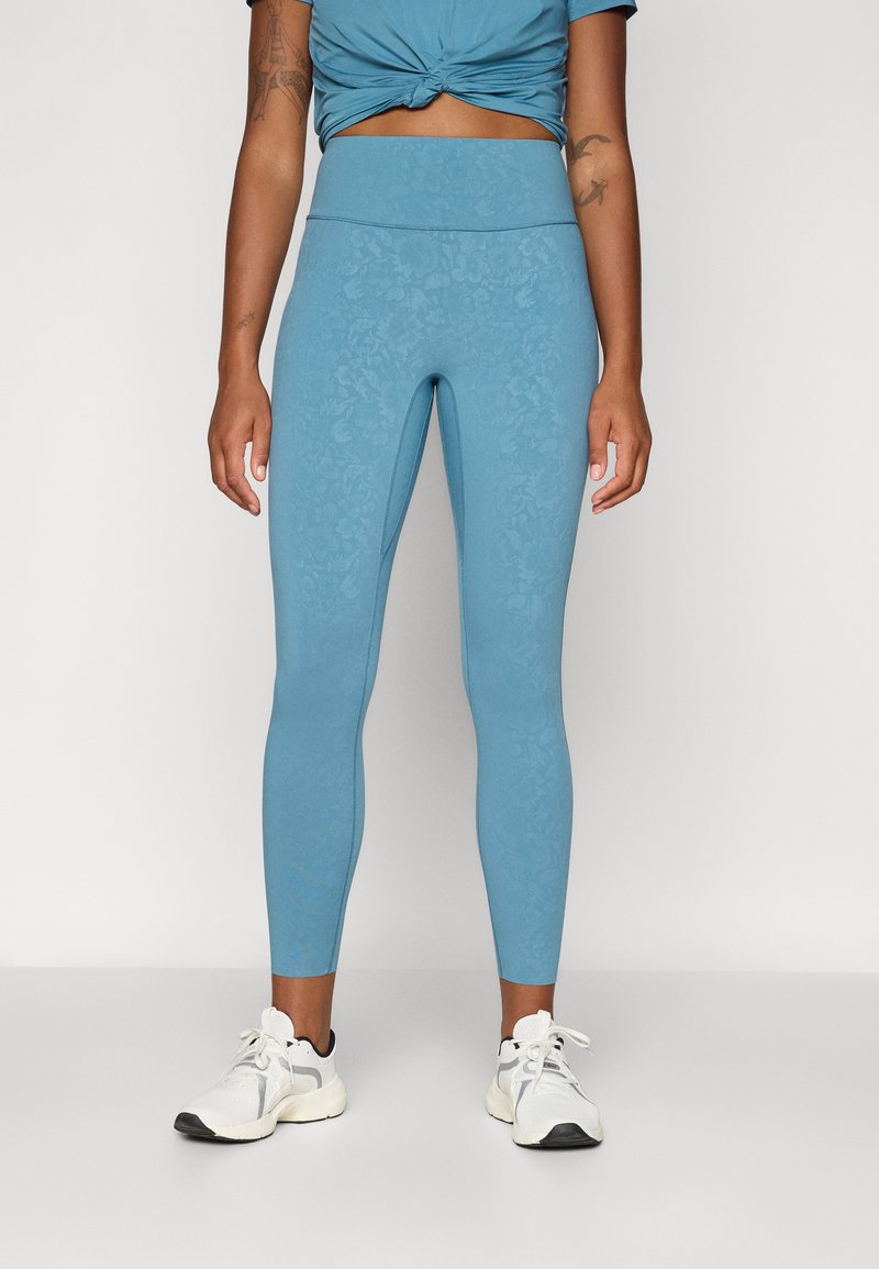 Lyseblå leggings med blomsterprint, høy midje og elastisk passform. Kombinert med hvite treningssko for et aktivt utseende.
