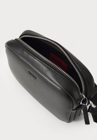 Borsa a tracolla in pelle nera con una texture liscia, zip argentata e un'etichetta interna con il marchio. Presenta una forma rettangolare compatta.
