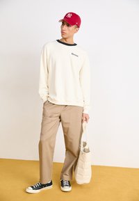 Béžová mikina s kontrastným čiernym výstrihom, krémové nohavice, čierne tenisky a červená čiapka, drží svetlobéžovú páperovú tašku na žltom podlahe.