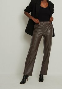 Kvinna i svart kavaj, svart topp, bruna högmidjade mockaskinnbyxor och svarta klackstövlar, står mot en plain bakgrund.