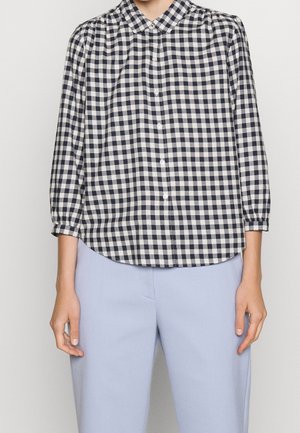 Osoba nosząca czarno-białą koszulę w kratę zapinaną na guziki z rękawami do łokcia oraz jasnoniebieskie spodnie na jednolitym tle.