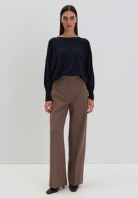 Marineblauer, oversized Pullover mit hohem Kragen und weichem Material. Braune, weit geschnittene Hose mit Bügelfalte. Dunkle Schuhe runden den Look ab.