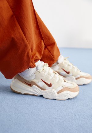 Fødder iført hvide og beige Nike-sneakers med røde swoosh-logoer, parret med orange bukser, stående på lyseblåt tæppe.