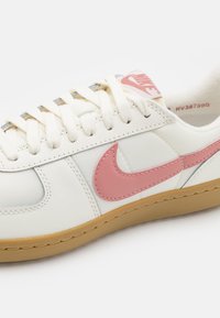 Baskets blanches Nike avec logo swoosh rose, lacets blancs et semelle en gomme, vues en gros plan de côté.