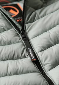 Chaqueta acolchada verde claro con cremallera completa, con textura acolchada, cremallera negra y una etiqueta con el texto "SUPERDRY" en blanco y naranja.