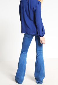 Blusa a maniche lunghe di colore blu navy con scollatura rotonda e chiusura a bottone sul retro, abbinata a jeans di denim blu a gamba larga.
