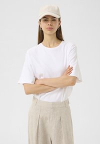 White cotton t-shirt with short sleeves, paired with high-waisted beige linen trousers; beige cap completes the outfit.