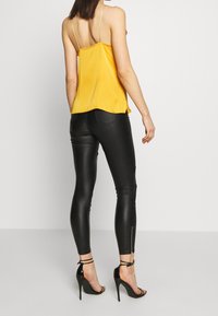 Žlutá saténová košilka s tenkými ramínky, kterou doplňují černé legíny z umělé kůže s zipy na kotnících a tvarovaným střihem.