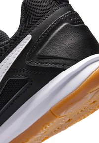 Κοντινή λήψη της φτέρνας ενός μαύρου αθλητικού παπουτσιού με δέρμα με υφή, λευκή μεσαία σόλα, εξωτερική σόλα από καουτσούκ χρώματος τσίχλας και λευκή ραμμένη διακοσμητική λεπτομέρεια στο πλέγμα επάνω μέρος.