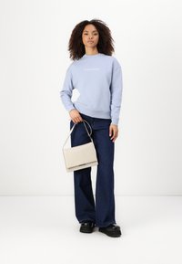 Sweatshirt azul claro com o logótipo "Calvin Klein", calças largas em azul escuro e uma bolsa creme. O modelo usa sapatos de plataforma pretos.
