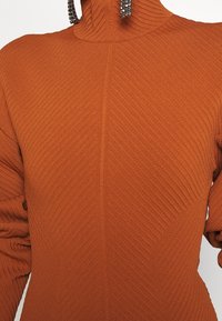 Gros plan d'un pull à col roulé en maille côtelée couleur rouille porté avec des boucles d'oreilles métalliques en chaîne visibles au niveau du cou.
