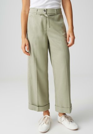 Femme portant un pantalon large vert clair avec les ourlets retroussés, des baskets blanches et un haut sans manches blanc, debout sur un fond gris.