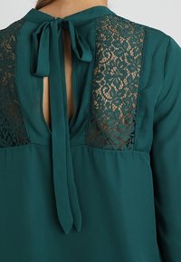 Turkos blus med spetsinsatser i ryggen, med en nyckelhålöppning och en rosettknut i halsen. Slät tygtextur.