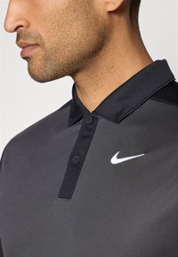 Nike polo krekls melnā krāsā ar teksturētu audumu, divām pogām un kontrastējošu baltu logotipu uz krūtīm. Īss apkakles dizains.