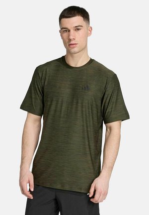 Hombre con una camiseta deportiva de manga corta verde oscuro con un pequeño logo en el pecho, de pie contra un fondo liso.