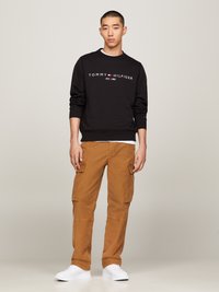Schwarzer Pullover mit weißem Text und Logo; beige Cargo-Hosen mit Seitentaschen; weiße Sneakers; einfarbiger Hintergrund.