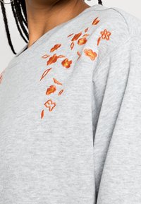 Sweatshirt gris avec broderie florale orange sur une épaule, en tissu texturé et à la coupe décontractée.