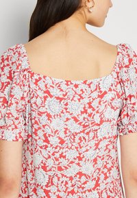 Červená květinová blůza s bílými a modrými botanickými vzory, čtveratý výstřih, krátké puff rukávy a měkká, texturovaná tkanina.