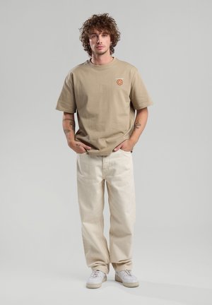 T-shirt en coton beige à manches courtes, avec un petit motif floral orange. Associé à un pantalon large beige clair et des baskets blanches.