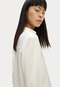 Blusa de seda blanca con cuello alto, mangas largas y una textura suave. Presenta plisados sutiles a lo largo de los hombros para un detalle adicional.