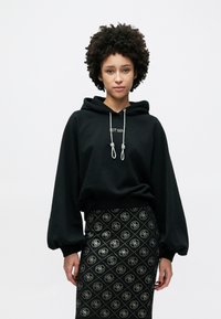 Μαύρο φούτερ με κουκούλα και κορδόνια, με μανίκια ραγκλάν, σε συνδυασμό με μαύρη φούστα με σχέδιο που επαναλαμβάνει το λογότυπο.