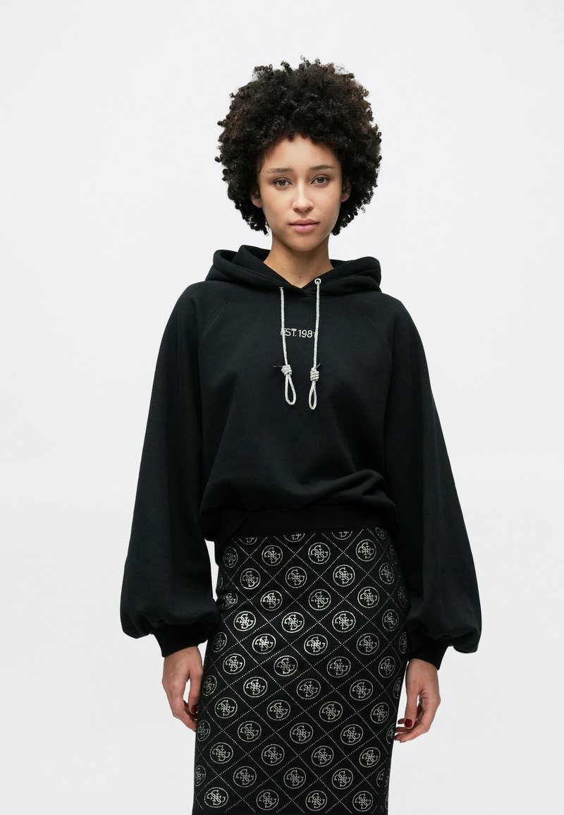 Μαύρο φούτερ με κουκούλα και κορδόνια, με μανίκια ραγκλάν, σε συνδυασμό με μαύρη φούστα με σχέδιο που επαναλαμβάνει το λογότυπο.