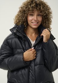 Chaqueta acolchada negra con acabado brillante, cuello alto y cremallera. Presenta secciones cosidas y botones a presión para el cierre.