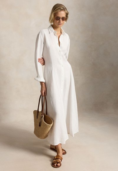 Γυναίκα που φοράει μακρύ λευκό φορεματάκι που κουμπώνει με κουμπιά, γυαλιά ηλίου, καφέ σανδάλια με λουράκι, κρατώντας υφαντή τσάντα ώμου με καφέ δερμάτινες λαβές.