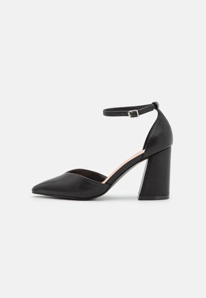 Chaussure à talon haut en cuir noir avec bout pointu, talon carré et bride de cheville ajustable avec boucle argentée sur fond blanc.