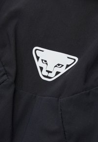 Tessuto nero con un logo triangolare che presenta un volto di animale stilizzato in bianco, con occhi acuti e elementi di design angolari.