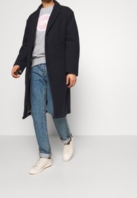 Cappotto in lana nera con felpa grigia scura, jeans in denim blu e sneakers bianche. Il cappotto presenta un colletto a rever e una vestibilità comoda.