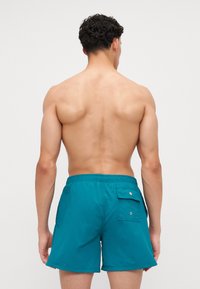 Shorts de bain turquoise en tissu léger, avec une poche au dos fermée par pression et une taille élastique.