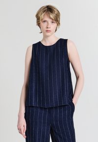 Jeune personne aux cheveux blonds courts portant un haut sans manches bleu marine et un pantalon à rayures assorti, debout devant un fond uni.