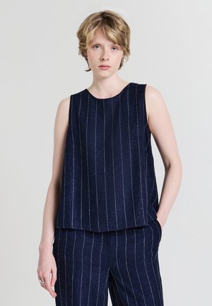 Jeune personne aux cheveux blonds courts portant un haut sans manches bleu marine et un pantalon à rayures assorti, debout devant un fond uni.