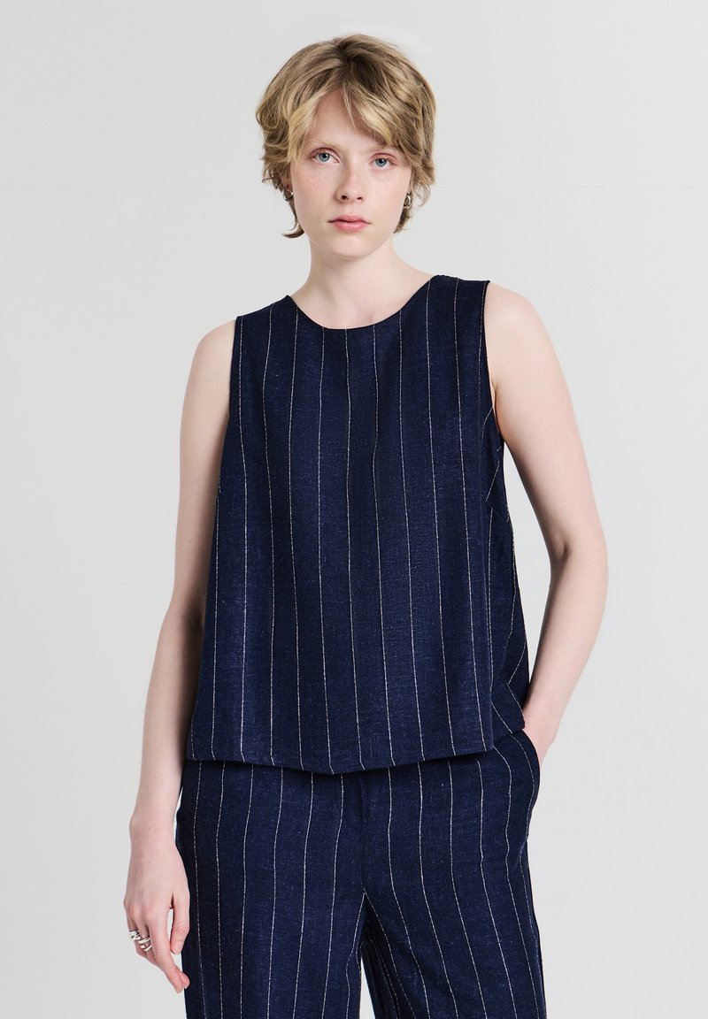 Jeune personne aux cheveux blonds courts portant un haut sans manches bleu marine et un pantalon à rayures assorti, debout devant un fond uni.