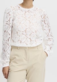 Blusa bianca in pizzo con motivi floreali, maniche lunghe a sbuffo e scollatura scallop, abbinata a pantaloni beige chiaro.