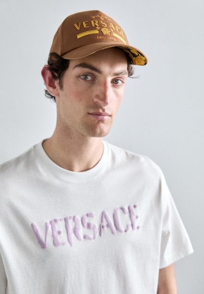 Căciulă maro cu logo auriu, subliniere modelată. Tricou alb cu inscripție ridicată "VERSACE" în culoare violetă. Fundal simplu.