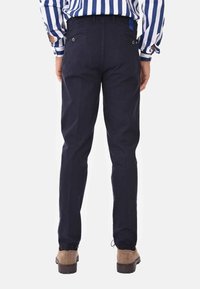 Pantaloni in cotone blu navy con vestibilità affusolata, polsini elasticizzati e tasche posteriori. Indossati con scarpe marroni e una camicia a righe visibile sopra.