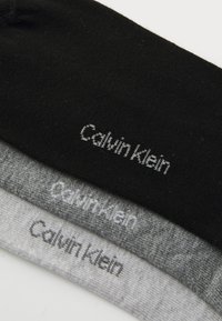 Tri par sock v črni, temno sivi in svetlo sivi barvi, z gladko bombažno teksturo in izvezenim logotipom "Calvin Klein" v sivi barvi.