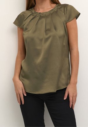 Woman wearing an olive green satin short sleeve blouse with pleats near the neckline and black pants against a white background.