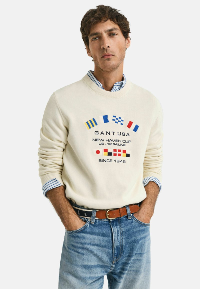 Crème sweater met geborduurde nautische vlaggen in blauw, rood en geel. Gecombineerd met lichtblauwe jeans en een brown riem. Colbert zichtbaar eronder.