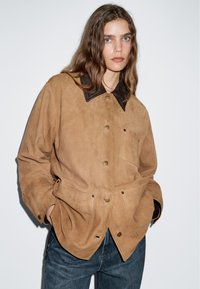 Giacca in suede marrone con colletto scuro, dotata di chiusura a bottoni, tasca sul petto e due tasche inferiori. Il tessuto ha una texture morbida.