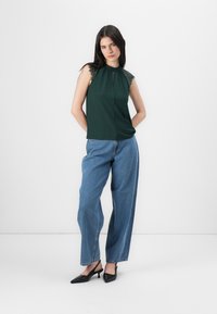 Una modelo posa con confianza en una blusa sin mangas verde con detalles de encaje, combinada con jeans de pierna ancha azules y tacones negros de punta.